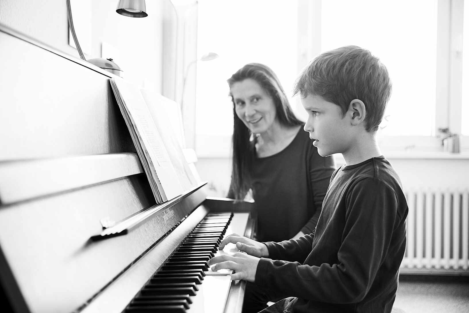 Kind spielt konzentriert Klavier. Lehrerin beobachtet den Jungen – ihren Klavierschüler.
